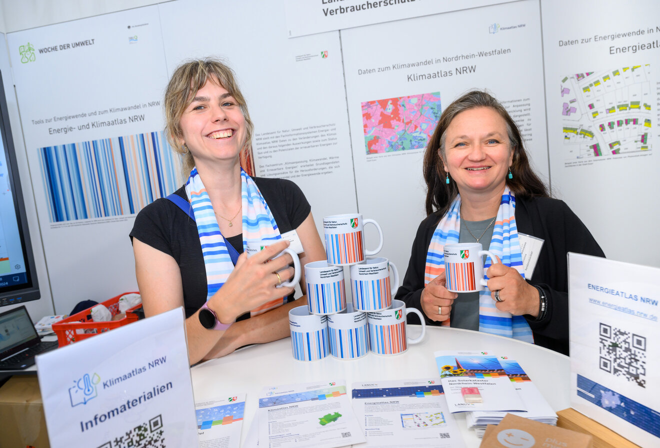 Foto vom Klimastand bei der Woche der Umwelt in Berlin. Zu sehen sind zwei Personen des Fachzentrum Klima. Auf dem Stehtisch im Vordergrund sind Tassen mit Warming Stripes von NRW zu sehen sowie verschiedene Flyer und Publikationen. Foto: David Ausserhofer / DBU